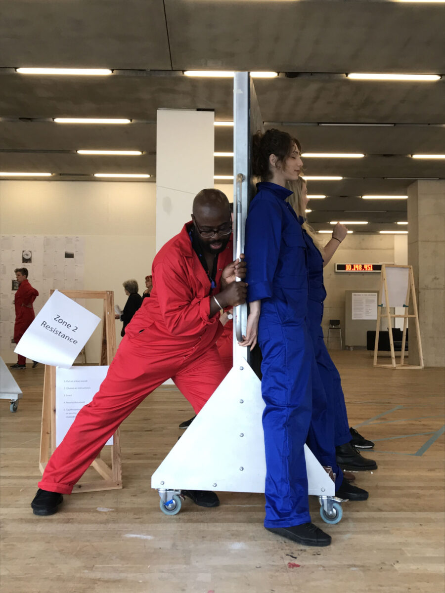 Guest artist and speaker Harold Offeh is invited by participants in blue overalls to be challenged over his superiority as a red-suited ‘expert’.